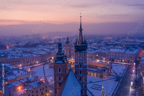 Main square Rynek of the Old Town of Krakow Poland in winter. St. Mary's Basilica Gothic church, Town Hall Tower, Cloth Hall, covered in snow at twilight with Christmas tree, decorations and lights.