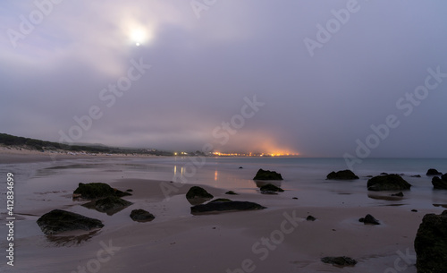 ocean and beach landscape with rocks under a stormy evening sky with full moon and village lights in the background