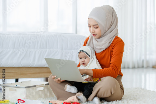 A cute and beautiful Asian Muslim mother work as an online seller working on laptop notebook computer and take care of her baby daughter in the same time