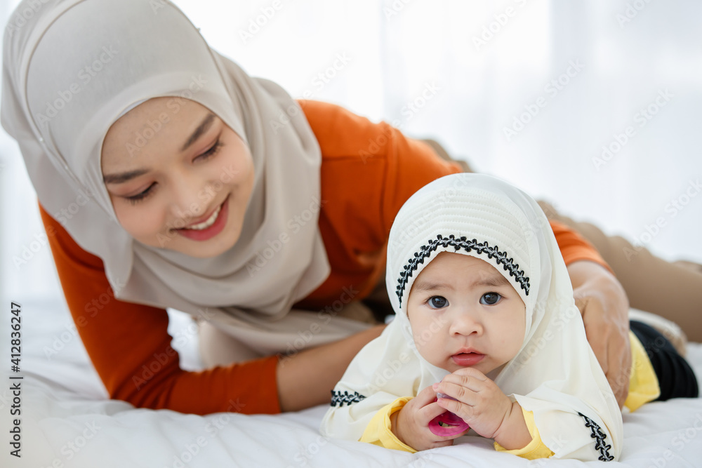 A cute and beautiful Asian Muslim in hijab dress playing with her daughter and playing for fun with pacifier on bed A cute and beautiful Asian Muslim in hijab dress playing with her daughter and playing for fun with pacifier on bed
