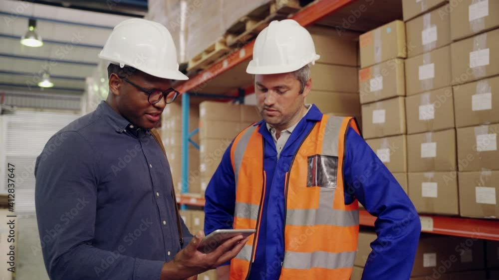 Smiling caucasian male warehouse worker and african manager looking at digital tablet discussing in storage factory Smiling caucasian male warehouse worker and african manager looking at digital tablet discussing in storage factory