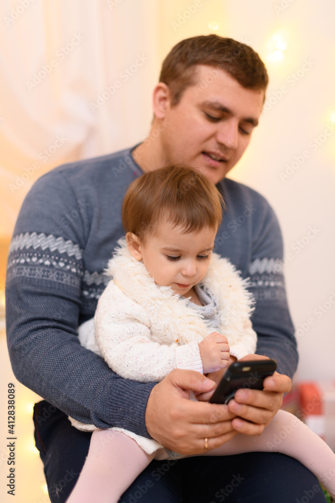 father and baby use smartphone, they play and watch device screen, home interior decorated with holiday lights and gifts father and baby use smartphone, they play and watch device screen, home interior decorated with holiday lights and gifts