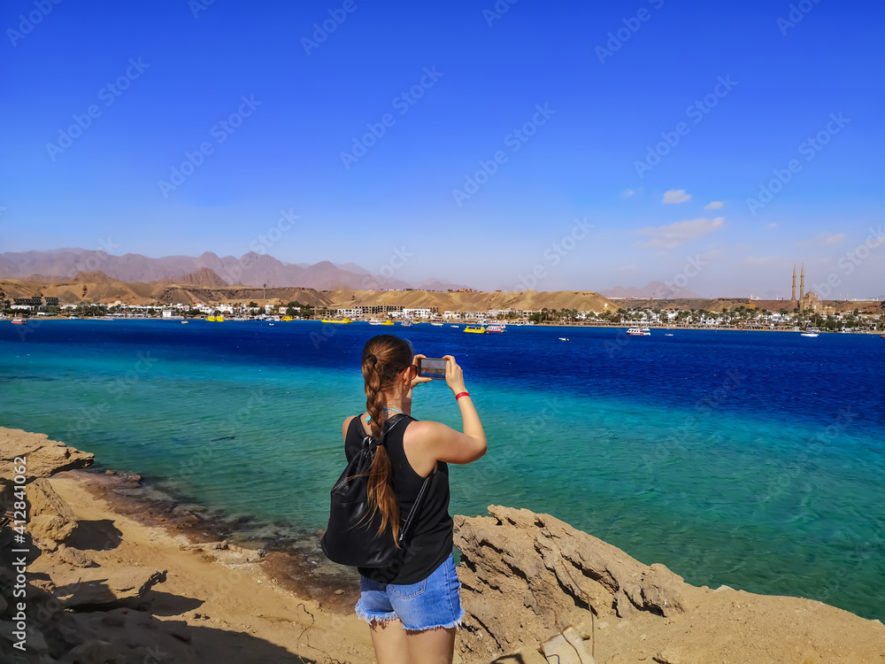 Young woman tourist takes pictures on a smartphone of the sea panorama of Sharm El Sheikh (Egypt). Exotic African landscape with an adult girl on the Red Sea shore Young woman tourist takes pictures on a smartphone of the sea panorama of Sharm El Sheikh (Egypt). Exotic African landscape with an adult girl on the Red Sea shore