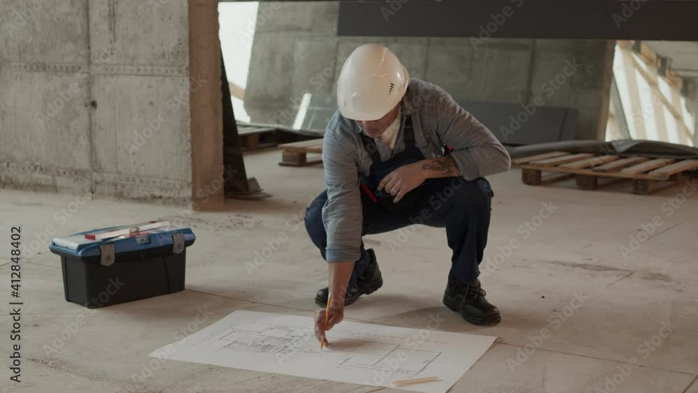 Full shot of Mixed-Race professional construction specialist wearing protective goggles and helmet in premises under renovation, correcting architectural blueprints of object on floor Full shot of Mixed-Race professional construction specialist wearing protective goggles and helmet in premises under renovation, correcting architectural blueprints of object on floor