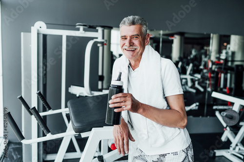Portrait of an older man holding a bottle of protein in his hands, he stands in the middle of the gym and cheerfully looks into the camera