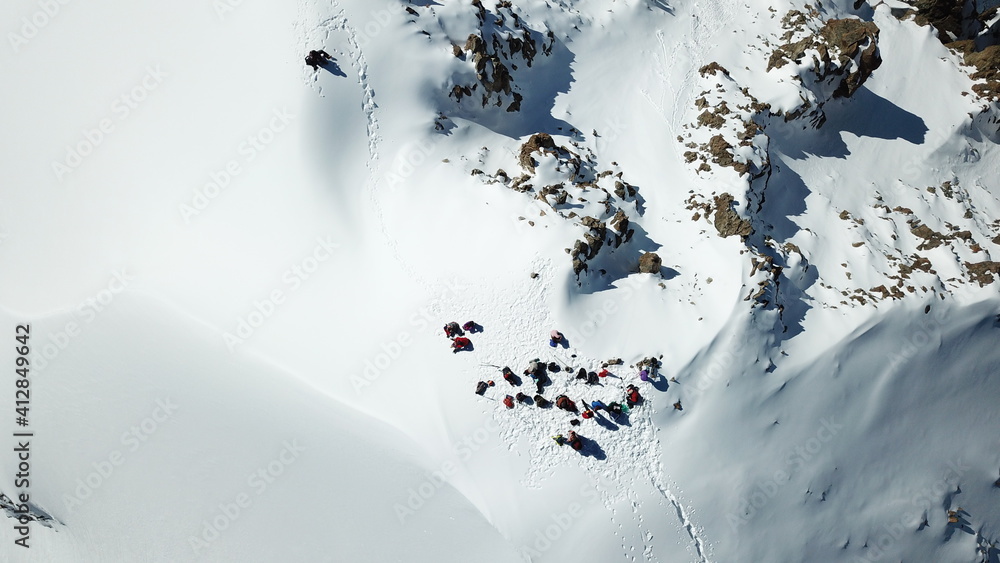 A group of climbers climb to the top of the peak. Dangerous, steep slope. Avalanche-prone area. Mountains covered with snow. Steep cliffs and large rocks. Above the mountains, top view from a drone.