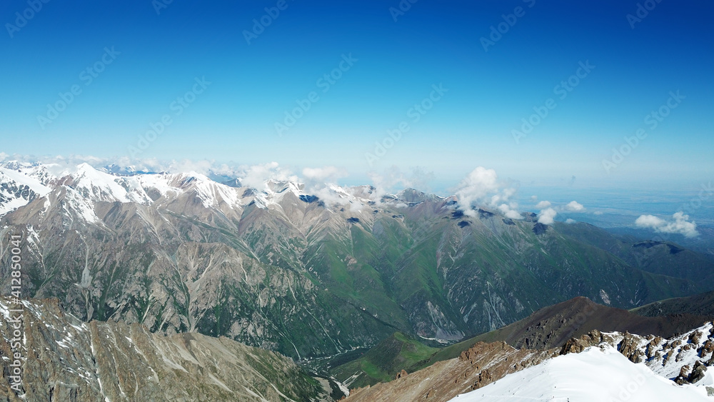 Snow-capped mountains and blue sky. View from a drone. Huge peaks covered with snow, steep cliffs. Blue, clear sky above the mountains. Flying over the peaks. Below you can see green trees and tarva.