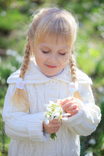Cute little girl considering a fresh bouquet of snowdrops. Spring time. Little girl in white walks in the forest
