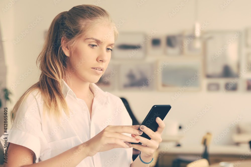 Focused beautiful blonde woman wearing white shirt, using smartphone standing in co-working space. Medium shot, copy space. Digital communication concept Focused beautiful blonde woman wearing white shirt, using smartphone standing in co-working space. Medium shot, copy space. Digital communication concept