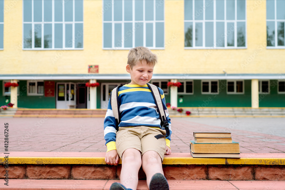 Portrait of Beautiful school boy looking happy outdoors at the day time. Sitting on the steps with books and a large school backpack school theme.