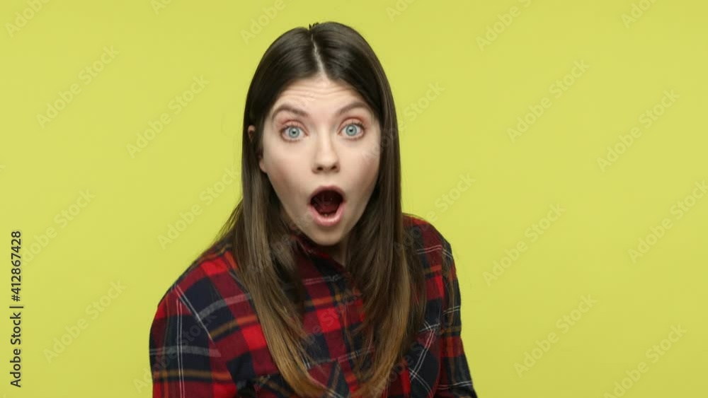 What? Extremely shocked surprised brunette woman in checkered shirt looking at camera with big eyes, widely opening mouth, expressing astonishment. Indoor studio shot isolated on yellow background What? Extremely shocked surprised brunette woman in checkered shirt looking at camera with big eyes, widely opening mouth, expressing astonishment. Indoor studio shot isolated on yellow background