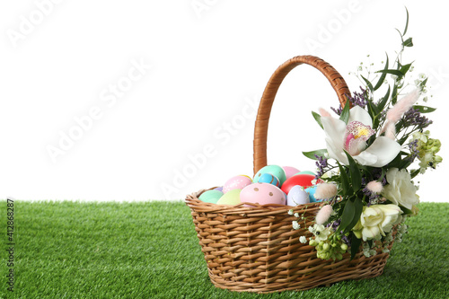 Wicker basket with Easter eggs and beautiful flowers on green grass against white background. Space for text