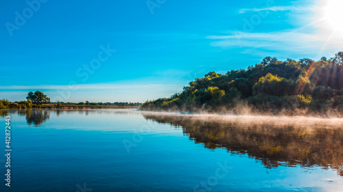 Aerial view of a beautiful summer landscape over river while dawn. Top view over river with a smooth water surface reflecting blue sky. Morning evaporation on a river while sunrise.