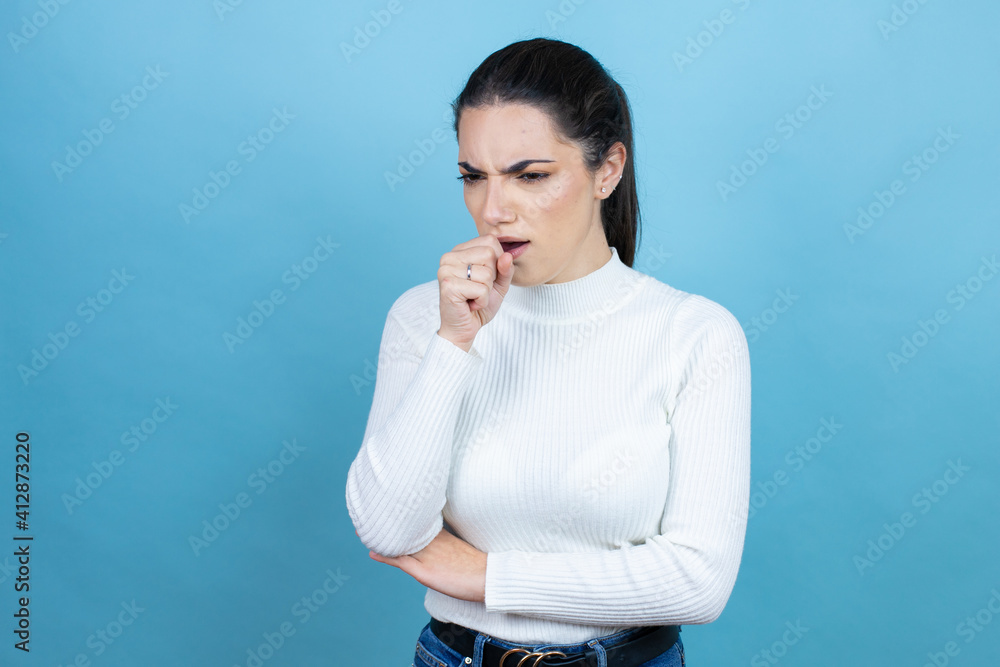 Young caucasian woman wearing white sweater over blue background with her hand to her mouth because she's coughing Young caucasian woman wearing white sweater over blue background with her hand to her mouth because she's coughing