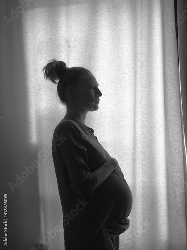 The silhouette of a pregnant girl standing by the window. Black and white photo.