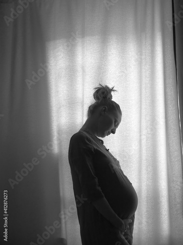 The silhouette of a pregnant girl standing by the window. Black and white photo.