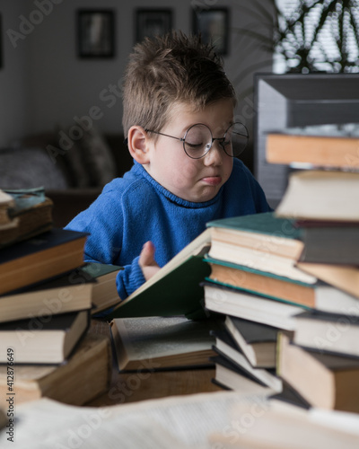 Five year old boy in glasses reading a book with a stack of books next to him. Smart intelligent preschool kid choosing books to borrow.