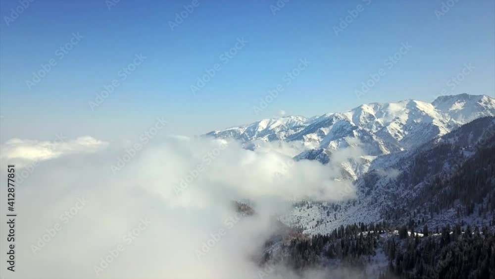 Flying among the clouds with a view of snowy mountains and forest. Huge clouds cover the trees on the hills of tmountain. All in the snow. White clouds and blue sky. Snow Kok Zhailau. Above mountains. Flying among the clouds with a view of snowy mountains and forest. Huge clouds cover the trees on the hills of tmountain. All in the snow. White clouds and blue sky. Snow Kok Zhailau. Above mountains.