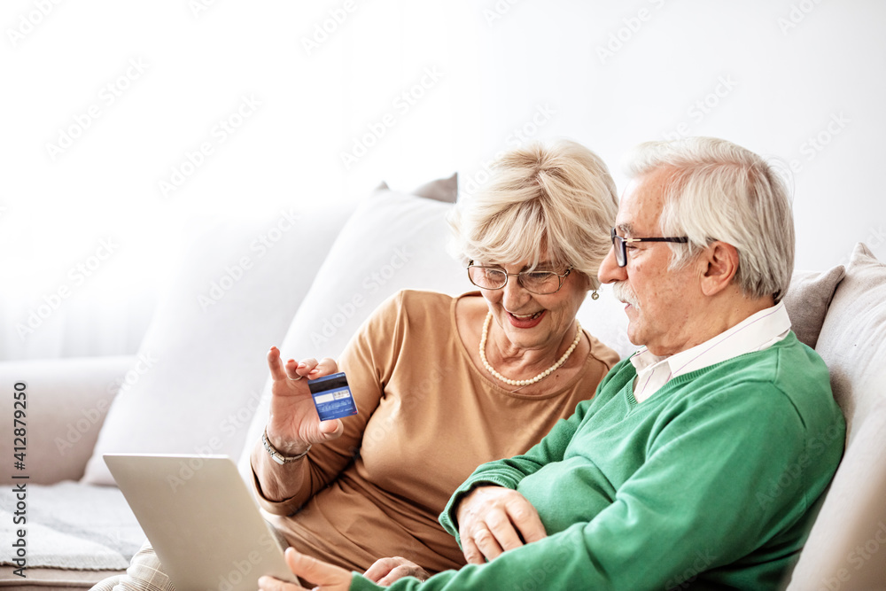 Senior couple at home sitting on sofa and shopping online on laptop. Senior husband and smiling wife paying bills online. Joyful middle aged couple doing online payment on digital tablet, copy space.