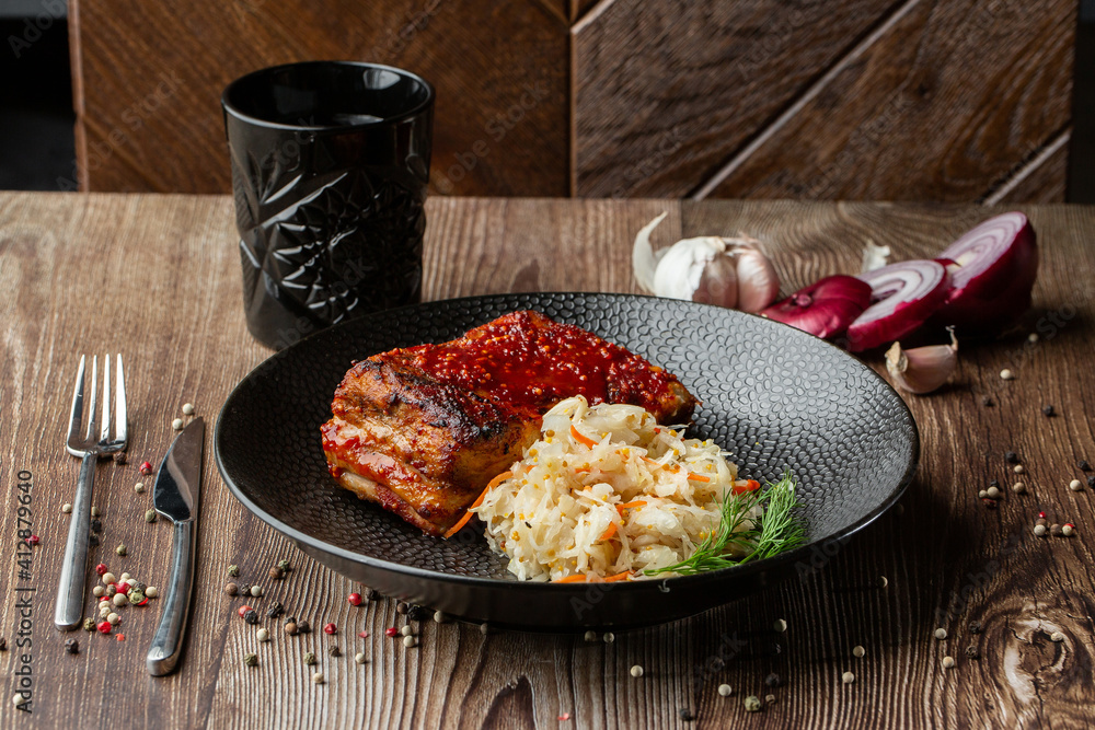 Baked meat served with red sauce, fermented cabbage and dill in black bowl. Close-up on a wooden background with a black glass, sliced onion, cloves of garlic, mix of pepper seeds and cutlery by side.