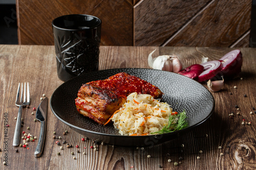 Obraz na płótnie Baked meat served with red sauce, fermented cabbage and dill in black bowl. Close-up on a wooden background with a black glass, sliced onion, cloves of garlic, mix of pepper seeds and cutlery by side. w salonie
