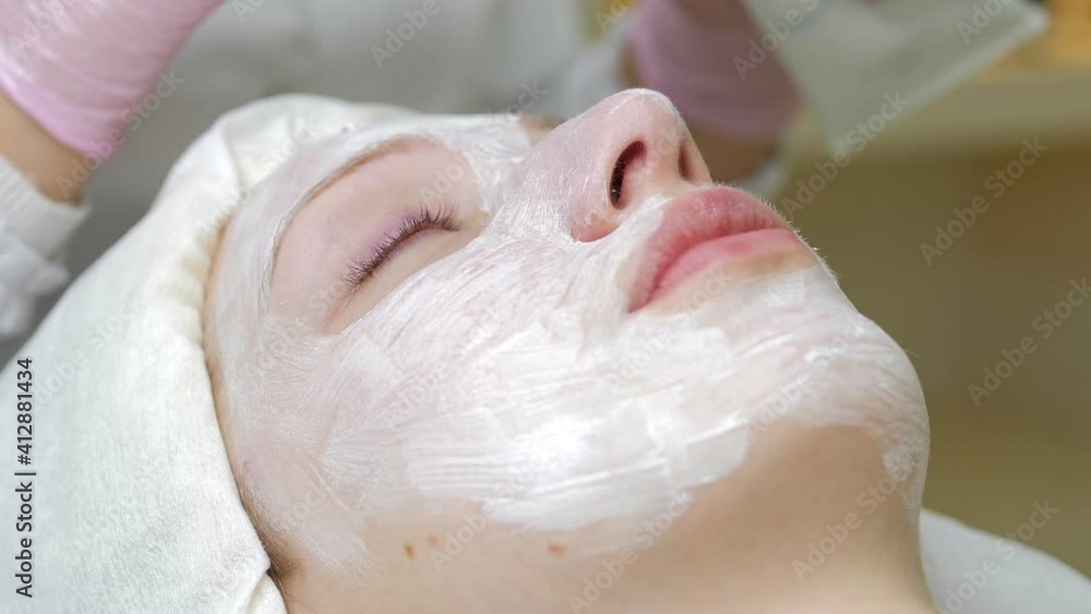 Experienced cosmetician is applying face mask on female face. Girl is lying and relaxing. Her eyes are closed with pleasure Experienced cosmetician is applying face mask on female face. Girl is lying and relaxing. Her eyes are closed with pleasure