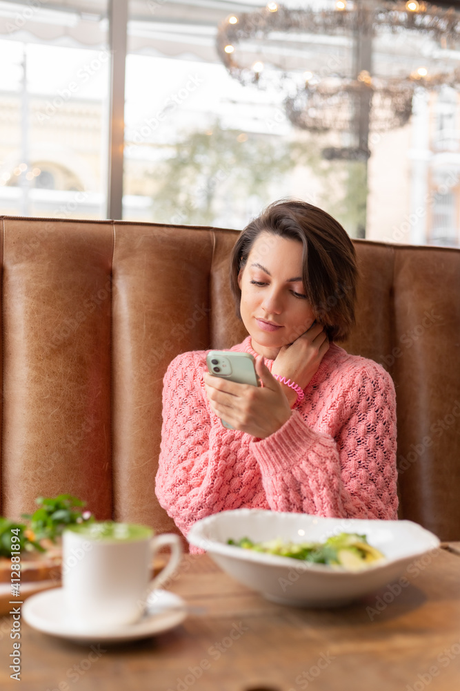 A woman in a restaurant in a warm sweater is looking at something on the phone, a delicious healthy breakfast and matcha latte on the table