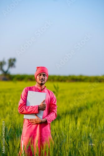 Young indian farmer holding in hand laptop at wheat field