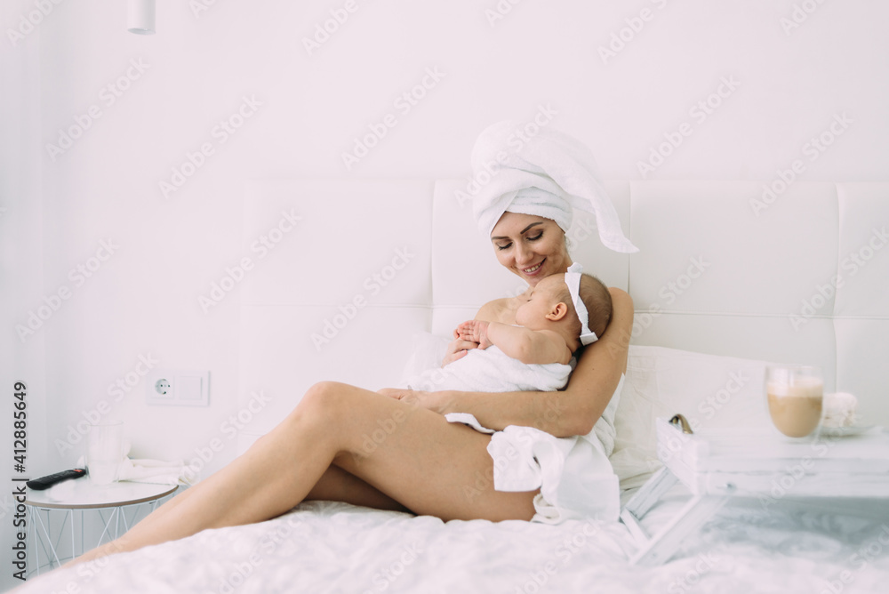 A Caucasian mother with a towel on her head sits on a white bed with bare feet after a shower and holds her little daughter in her arms. A Caucasian mother with a towel on her head sits on a white bed with bare feet after a shower and holds her little daughter in her arms.