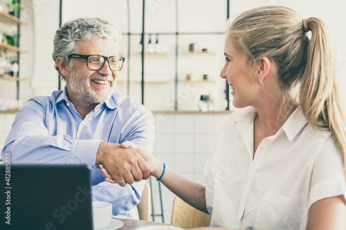 Businessman meeting and shaking hands with insurance agent at co-working, sitting at open laptop, smiling, discussing agreement. Medium shot,. Handshake or cooperation concept