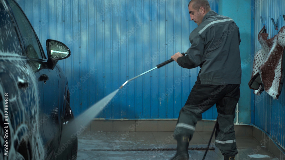 A Caucasian man works in a car wash service. A man in a worker's uniform washes the car thoroughly by spraying it with a water pistol. A Caucasian man works in a car wash service. A man in a worker's uniform washes the car thoroughly by spraying it with a water pistol.
