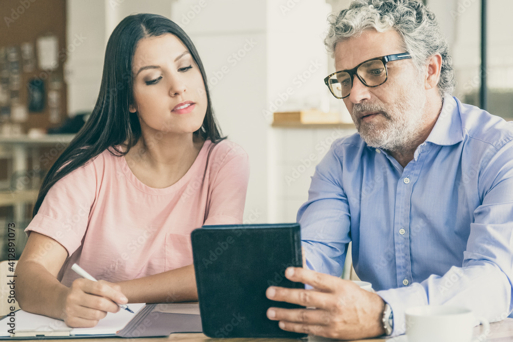 Serious mature male professional showing content on tablet to young colleague. Woman looking at screen. Medium shot, side view. Digital communication concept Serious mature male professional showing content on tablet to young colleague. Woman looking at screen. Medium shot, side view. Digital communication concept