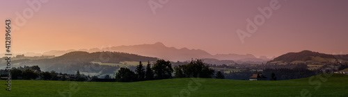 Fotografie Panoramic shot of a greenfield on a mountain landscape background during sunt