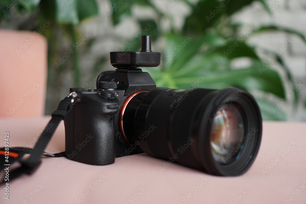 Black camera with a lens on a pink background and a blurred background of green leaves. No visible logos and brands