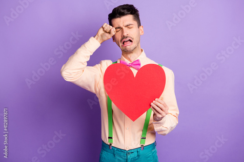 Photo of unhappy lonely young guy wear yellow shirt holding big paper red heart crying tears isolated violet color background © deagreez
