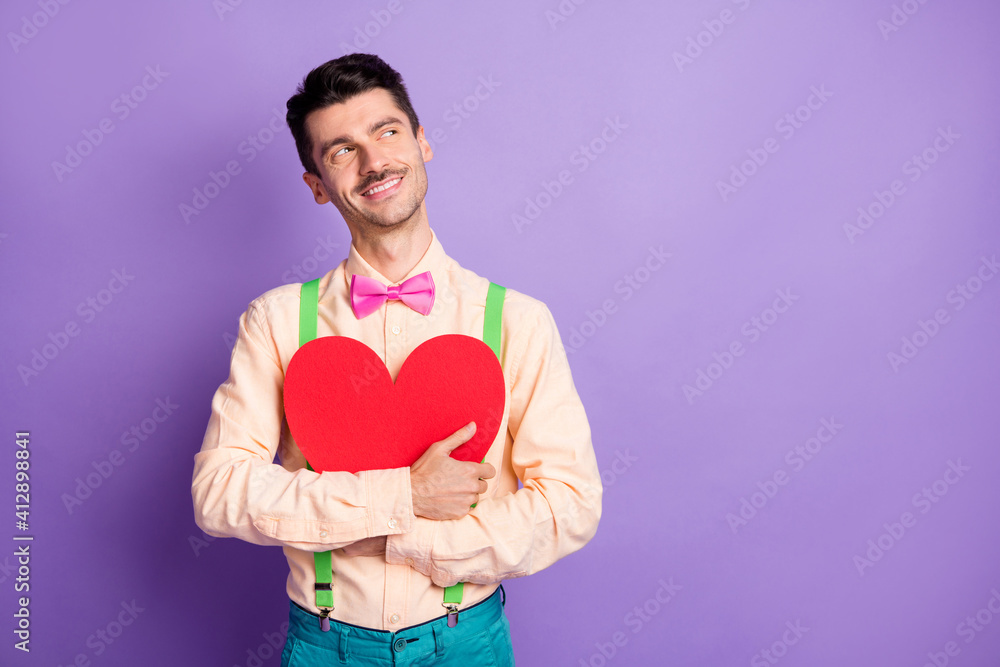 Photo of cute sweet young guy wear yellow shirt embracing big paper red heart looking empty space isolated violet color background Photo of cute sweet young guy wear yellow shirt embracing big paper red heart looking empty space isolated violet color background
