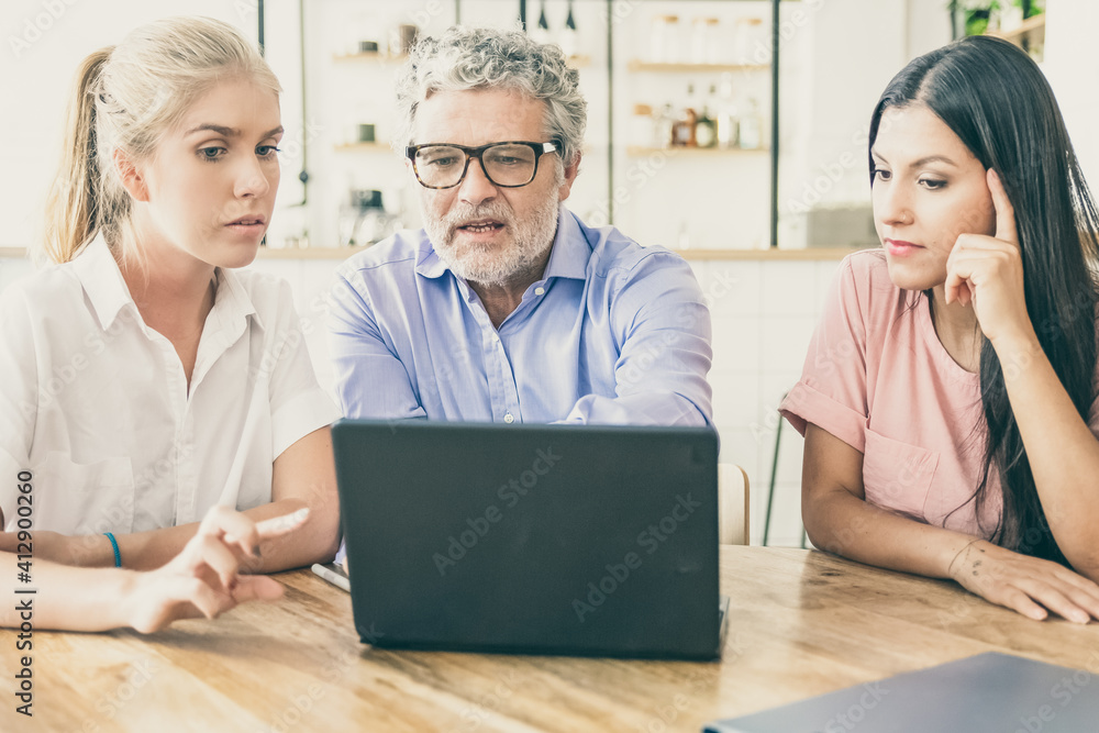 Focused pensive young woman and mature man meeting with female professional, watching and discussing content on laptop. Man pointing finger at display. Consulting or cooperation concept Focused pensive young woman and mature man meeting with female professional, watching and discussing content on laptop. Man pointing finger at display. Consulting or cooperation concept