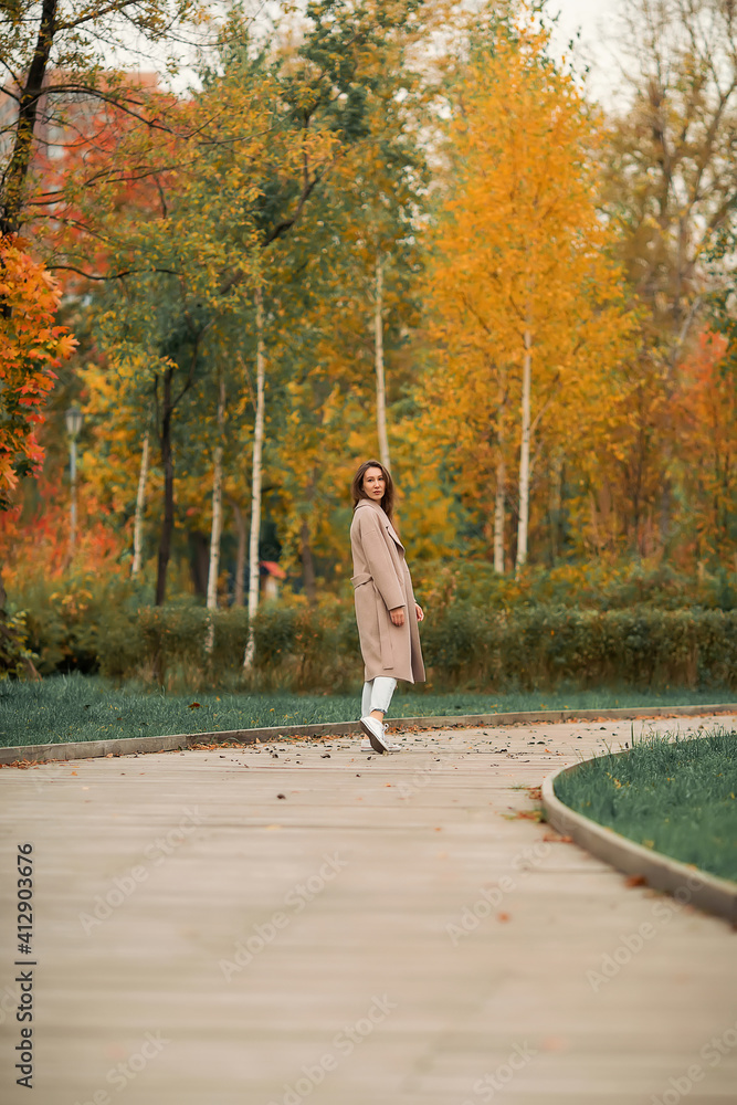 Fototapeta premium A girl with a Euro-Asian appearance in a pink coat and blue jeans with holes walks in an autumn park