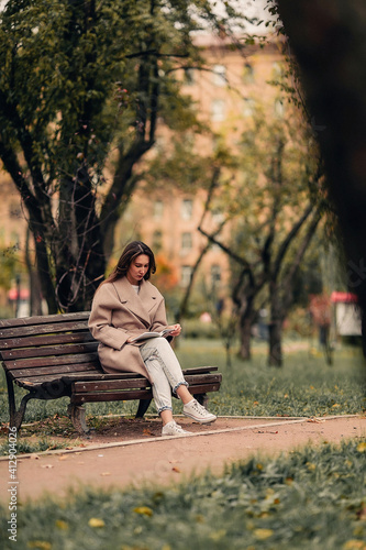 A Caucasian girl in a beige coat and blue jeans sits on a bench in a park in autumn and reads a book
