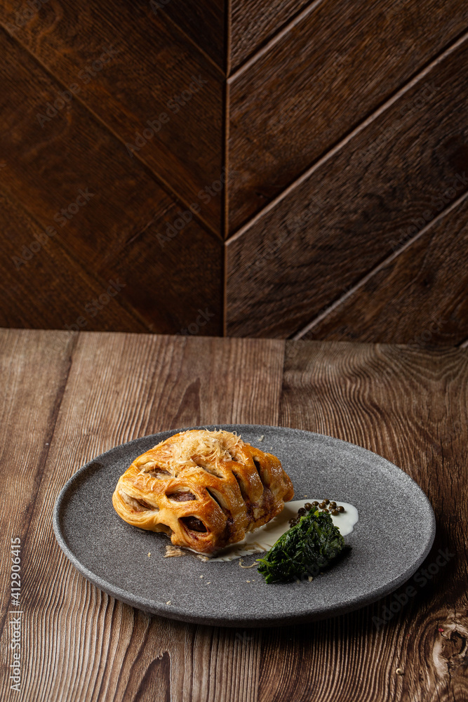 Puff pastry with meat and cheese on top served with mashed spinach and white sauce. Close-up of the dish on a grey plate isolated on wooden background. Vertical orientation, side view, copy space.