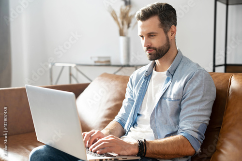 Clever freelancer using laptop for remote work from home. A handsome bearded guy websurfing, sends and answering emails, develops software sittin on the couch at home. Intelligent man works remotely