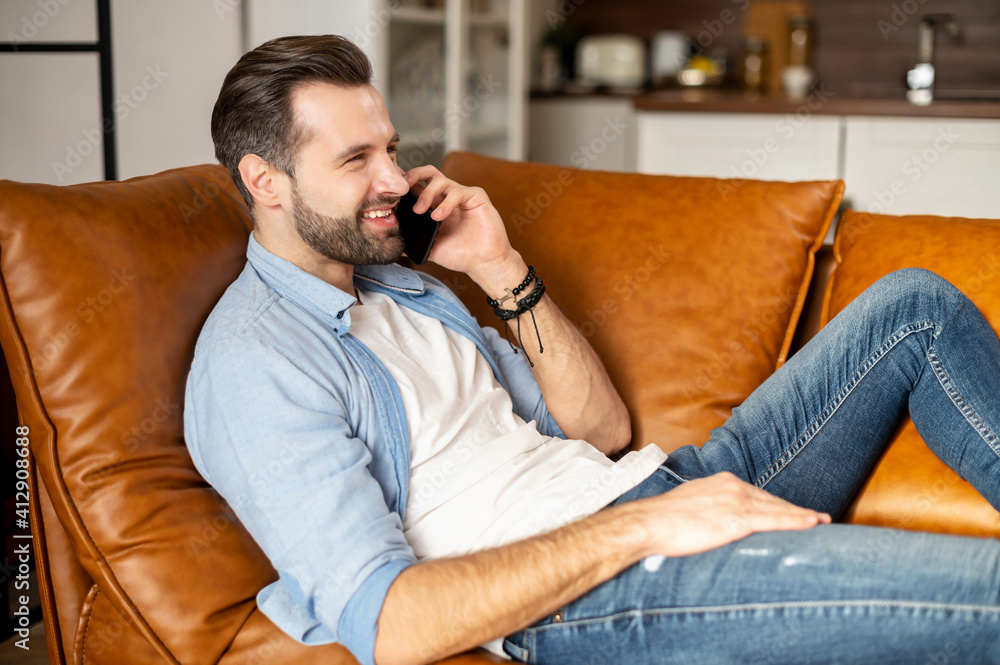 Side view an attractive bearded hipster talks by the smartphone lying on the comfortable couch, smiling young man has phone conversation at home