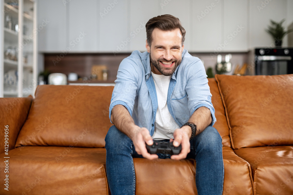 Handsome excited young guy spends leisure time playing video game, gambling guy with a joystick sitting on the couch, play on console at home