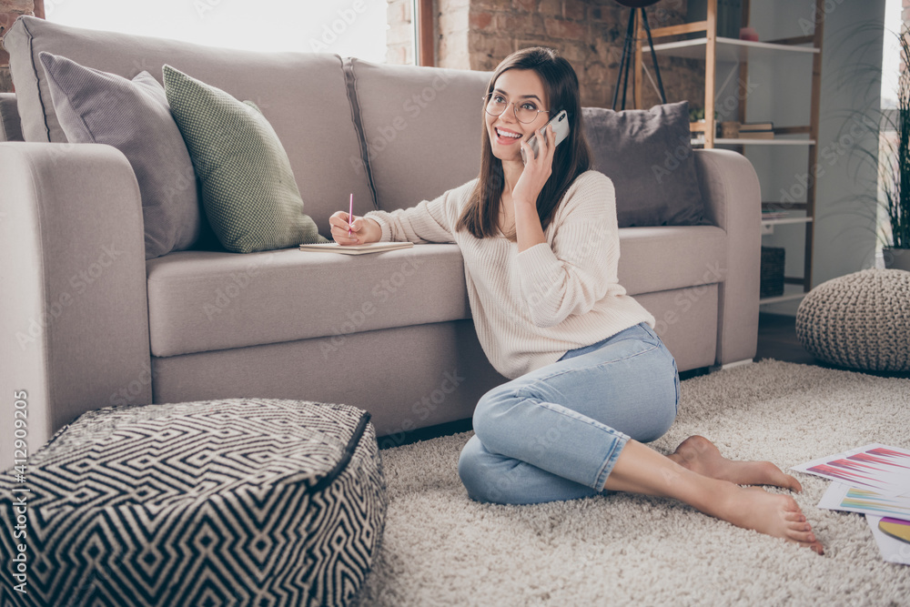 Full length photo of positive charming girl speak phone write note good mood sit home floor inside house indoors Full length photo of positive charming girl speak phone write note good mood sit home floor inside house indoors