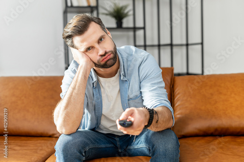 Fotografía Handsome bored young man holding tv remote control, watching boring TV shows, sw