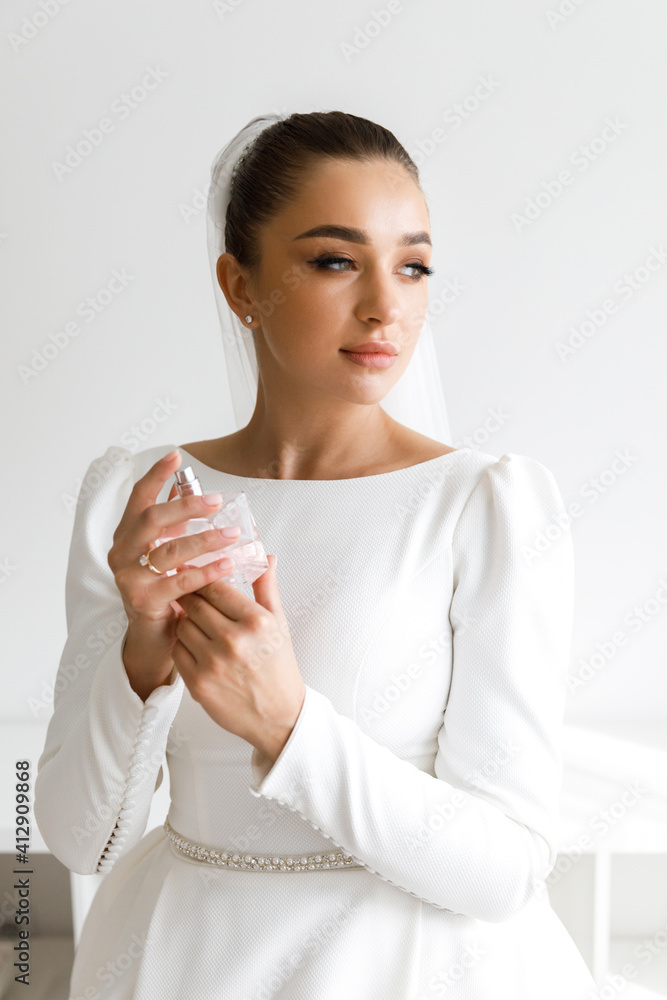 Beautiful luxurious bride holding a bottle of perfume. Morning of the bride in a white dress and a veil in the room Beautiful luxurious bride holding a bottle of perfume. Morning of the bride in a white dress and a veil in the room