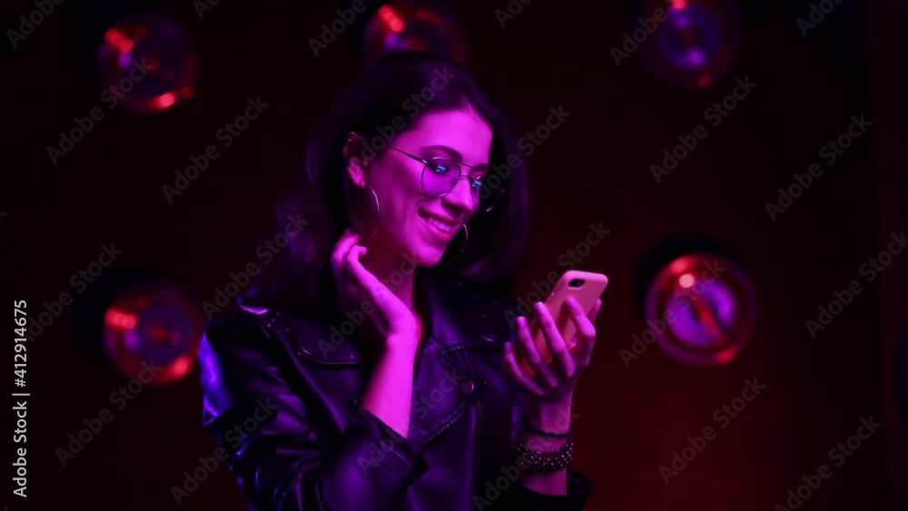Beautiful brunette girl in club atmosphere. Happy female in sunglasses standing in room in the pink and blue neon light, texting on cellphone and smiling. Close up. Slow motion. Beautiful brunette girl in club atmosphere. Happy female in sunglasses standing in room in the pink and blue neon light, texting on cellphone and smiling. Close up. Slow motion.