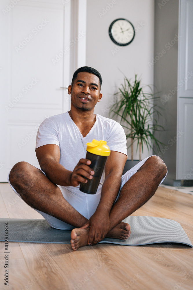 Front view of cheerful muscular African-American man sitting cross-legged relaxing on exercise mat after workout and holding proposing bottle of fresh mineral water at domestic room.