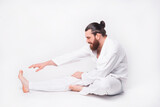 Photo of young bearded man wearing taekwondo uniform making stretching over white background.