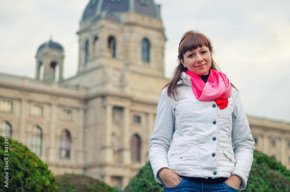 Young girl traveler with white jacket and red scarf looking at camera and posing, Kunsthistorisches Museum of Art History or Fine Arts in Vienna city historical centre background, vacation in Austria